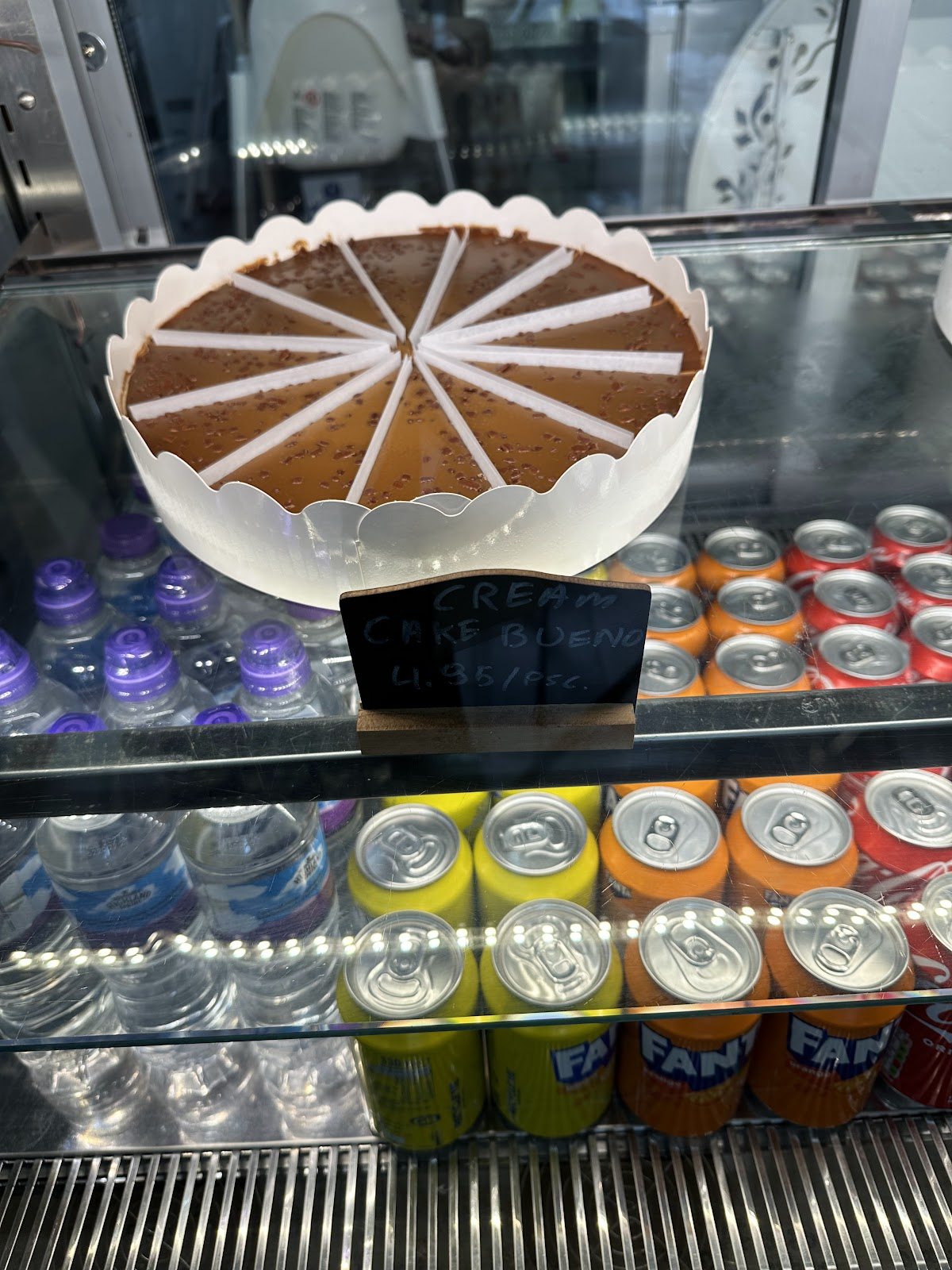 A glass cabinet with a whole chocolate-topped cream cake cut into slices, above chilled drinks.