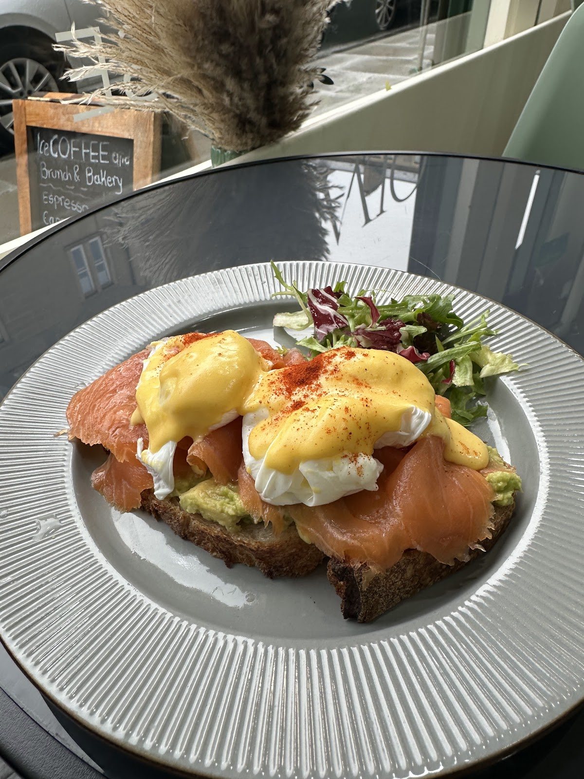 Eggs royale: smoked salmon and poached eggs under hollandaise and paprika on sourdough with smashed avocado and a leaf salad, on a ridged grey plate at a glass table.