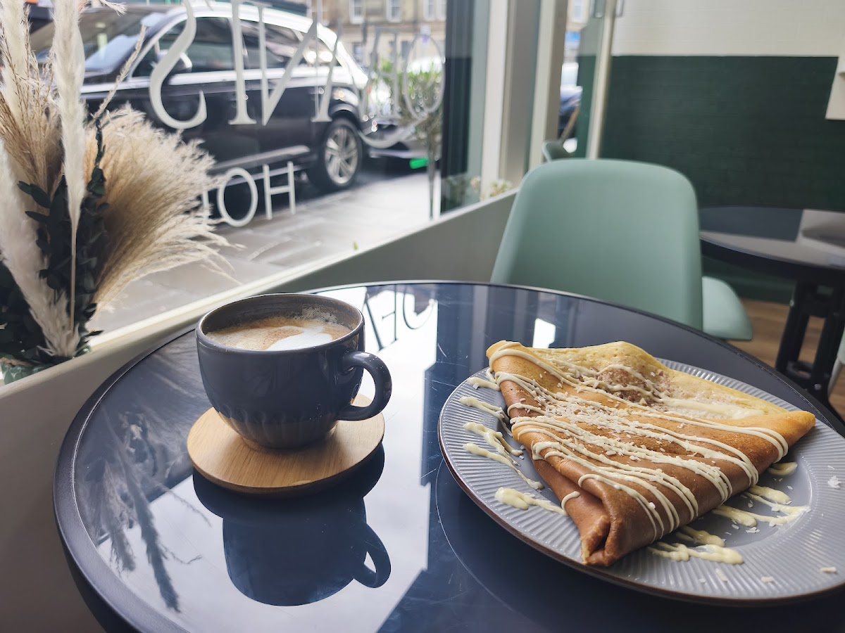 A crêpe with a white chocolate drizzle and a cup of coffee on a glass table by the window, with pampas grass in the corner.